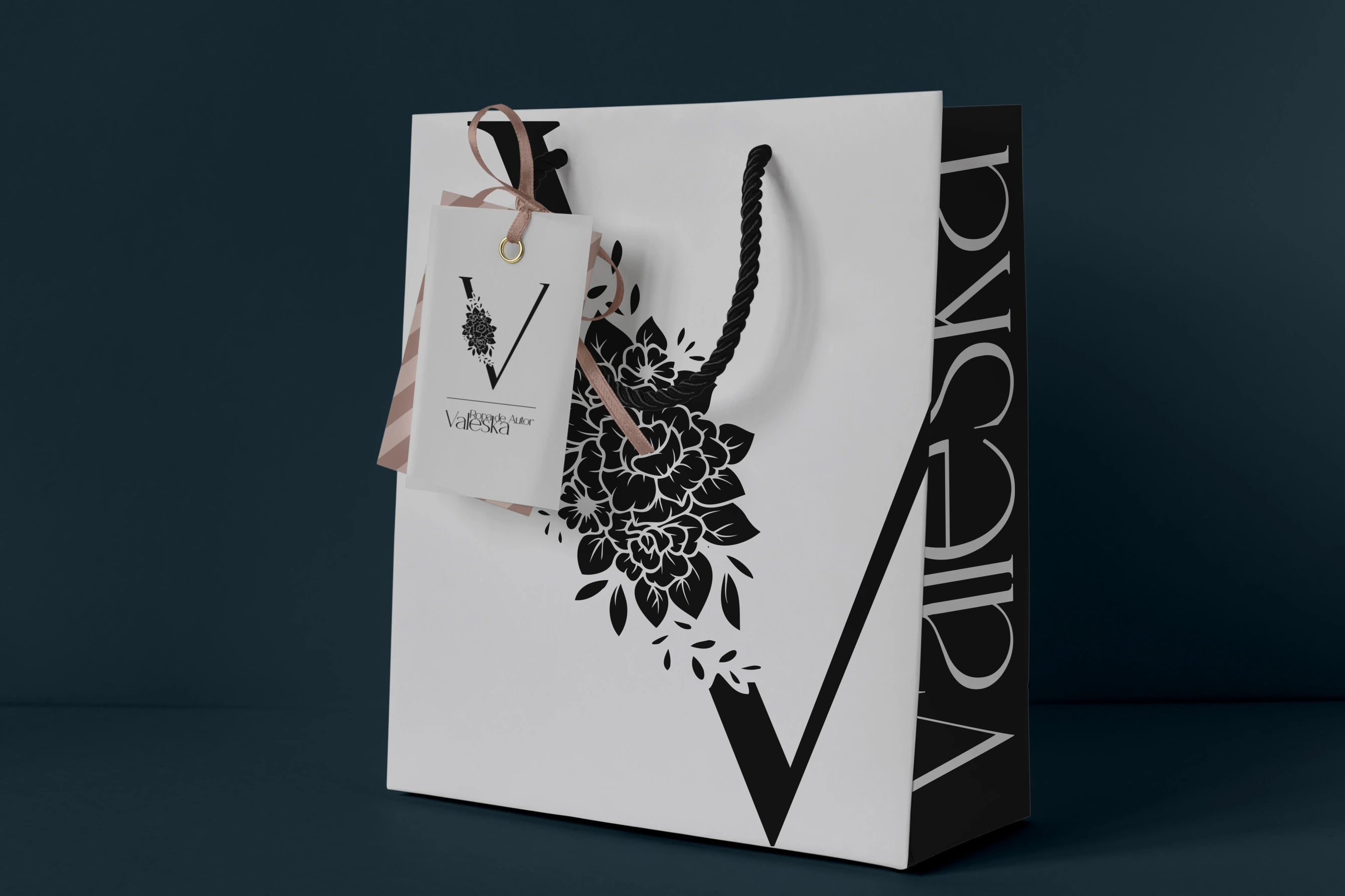Diseño de la bolsa de compras de la marca. Con el frente de fondo blanco mostrando una rosa negra y la letra V, y costados negros con el nombre de la marca.